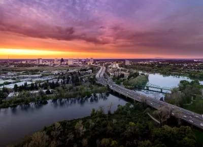Aerial view of freeway crossing river in Sacramento, California with colorful sunset sky in background.