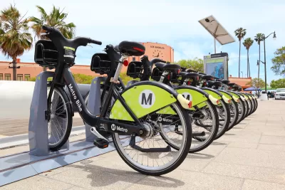 Row of black and green Los Angeles Metro bike share bikes at station with red building with clock and'Los Angeles' lettering in background.