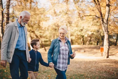 Grandparents walking with young grandchild through park with fall foliage.