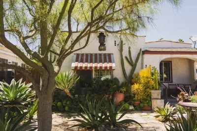 Front of an Spanish style bungalow with striped window awnings and a tree and yard landscaped with cacti.