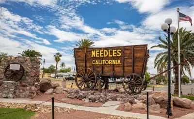 Sign for Needles, California installed on wooden wagon with residential neighborhood visible in background.