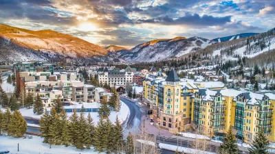 Aerial view of Vail, Colorado ski resort town with multi-story buildings in foreground and snowy mountains in background.