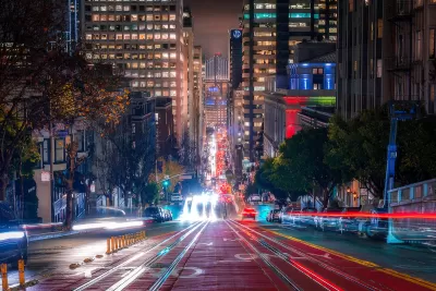 Blurred lights of traffic in motion on San Francisco street with high-rise buildings and Bay Bridge visible in background at night.
