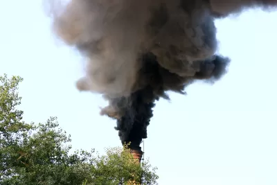 A factory smoke stack billows out black smoke into a blue sky.