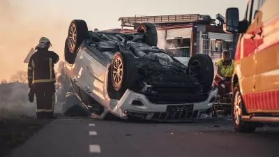 Car crash scene with overturned car, emergency vehicles, and firefighter standing at left.