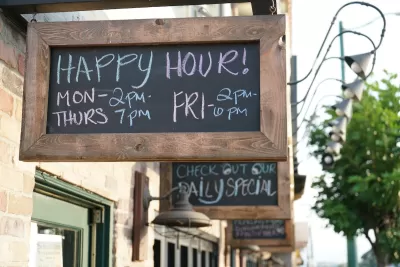 Close-up of rustic wood-framed chalkboard sign with happy hours listed above green restaurant doorframe on small commercial street.