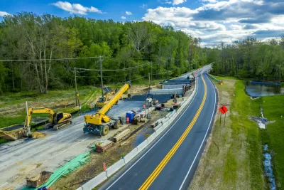 Aerial view of road construction with yellow heavy equipment parked next to two-lane asphalt road against green forest and blue sky