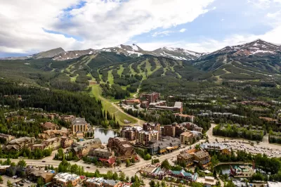 An aerial view of the town of Breckenridge, Colorado, in the foreground with lots of businesses, houses, and a ski resort, with green treed mountains in the backround and taller snow-capped mountains in the distance.