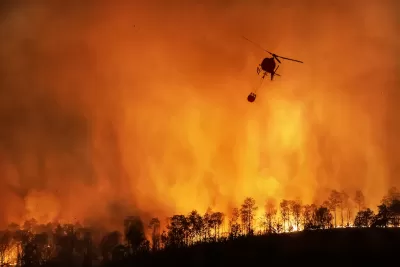 Firefighting helicopter sillhouetted against orange nighttime wildfire in California with trees visible at bottom of image.