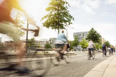 Line of five cyclists bike away from camera in wide protected bike lane on the right side of a street, with low-rise residential buildings in the background.