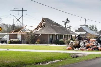 Exterior side view of a single-story brick home missing back half of roof and a debris pile in the front yard.
