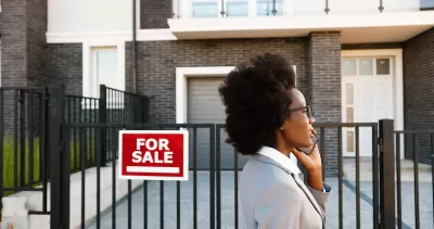Woman realtor or home buyer client wearing blazer walking past urban condo with red and white For Sale sign while talking on phone.