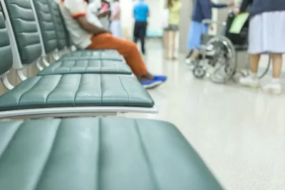Close-up of blurred empty vinyl chairs with person sitting in background and wheelchair visible in hospital waiting room.