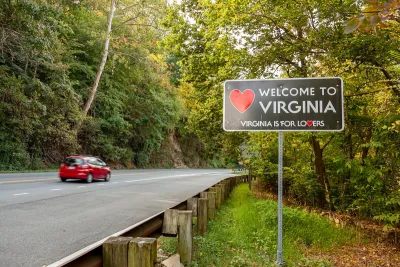 Small red car driving on forested road passing "Welcome to Virginia" sign.