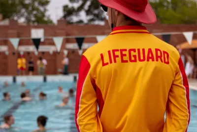 View from behind lifeguard standing in front of crowded public swimming pool wearing yellow shirt with LIFEGUARD in red lettering and red sun hat