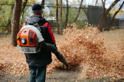 Man in black and red jacket using backpack-mounted leaf blower on large pile of brown leaves.
