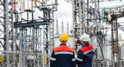Two men in white and yellow hard hats and matching jackets looking up at power infrastructure in outdoor electrical substation or plant.