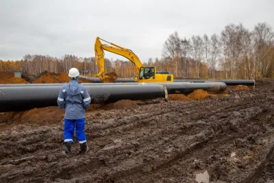 Worker in hard hat stands in front of oil pipeline under construction with yellow heavy equipment.