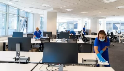 Professional cleaners wearing medical masks and gloves cleaning desks in large empty office.