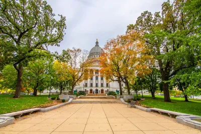 Green trees, some with fall colors, line a walkway in front of the Wisconsin state capitol dome in Madison, WI.