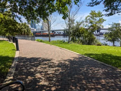 Brick path through an urban park shaded by trees with a river on the righhand side and high-rise buildings in the distance.