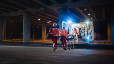Three EMTs in orange uniforms putting gurney with person onto ambulance at night under freeway overpass.