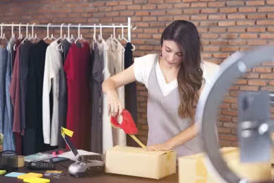 Woman taping up a brown paper package with rack of clothing in background.