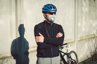 Male cyclists wearing helmet, sunglasses, and N95 mask stands with arms crossed in front of his bike