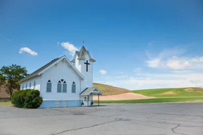 Small white wooden church at edge of large concrete lot with rolling hills or golf course in background.