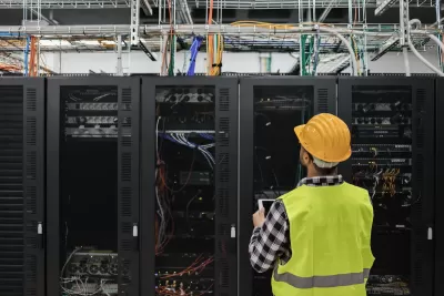 Worker in yellow safety vest and hard hat looks up at servers in data center.