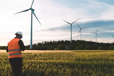 Technician in orange vest and white hard hat standing in field with row of wind turbines.