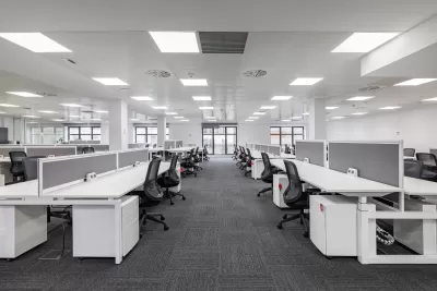 Large empty open-plan office with rows of white desks and black chairs under bright fluorescent lighting