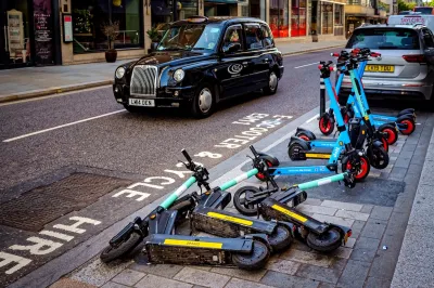 Group of e-scooters messily parked on street in London with black cab in background.