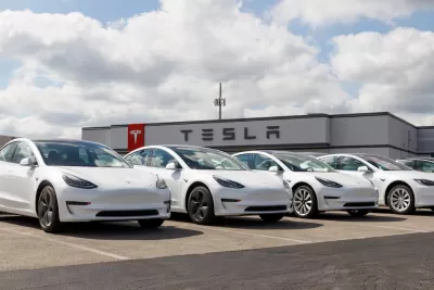 Row of white Tesla electric cars in parking lot with dealership building with Tesla logo in background.