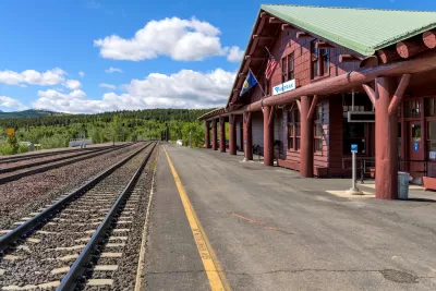 Rail tracks on the left, rustic log-built train station painted reddish brown with a green metal roof and concrete platform on the right, evergreen forest and bright blue sky with fluffy white clouds in the background.