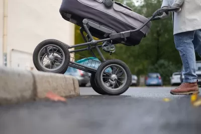 Close-up of bottom half of stroller being pushed onto sidewalk with no curb cut by person in jeans and brown shoes.