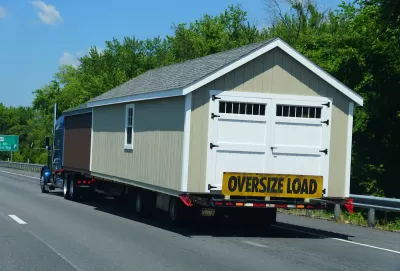 Tan modular building with white swinging barn/garage door being transported on freeway with yellow "Oversize Load" banner across back.