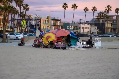 Colorful tents built to shelter unhoused people on beach at Venice Beach, California with apartment buildings and palm trees in background against a purple sunset sky