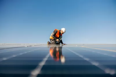Worker in safety vest and white hard hat installing rooftop solar panel.
