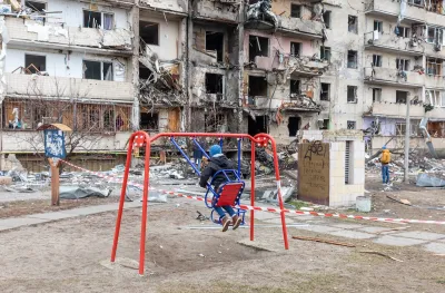 Child on playground swing in front of bombed building in Ukraine.