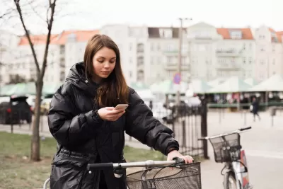 Woman stands next to a bike with one hand on handlebar holding her mobile phone.