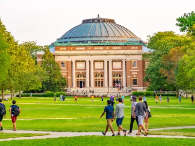 College students walking on green lawn with neoclassical red brick domed building in background on University of Illinois campus.