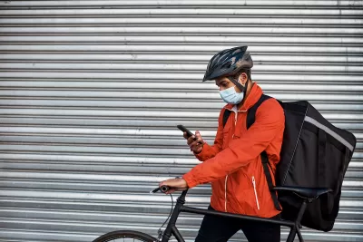 Man in orange jacket wearing surgical mask and bike helmet looks at phone while wearing delivery backpack and holding bicycle