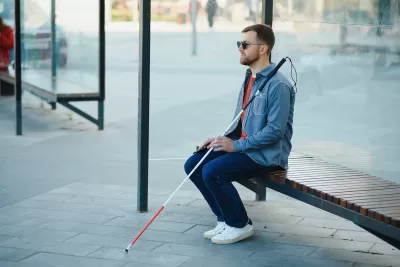 Man with sunglasses and cane sits on bus stop shelter bench.