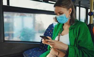 Woman in green sweater wearing light blue medical mask sitting on bus looking at her phone.