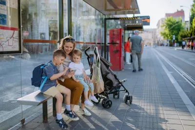 Woman and two children sit on bench at public transit stop waiting for tram with stroller next to them.