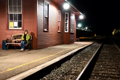 Womanin yellow jacket sitting on bench at outdoor train station with red wood depot building at night.