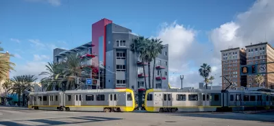 Two silver trains leaving station in Long Beach, California in front of mid-rise residential building.