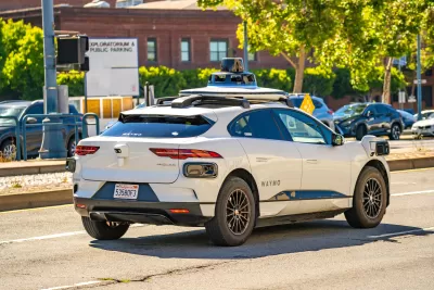 White Waymo self-driving car with roof-mounted camera driving on San Francisco street with Exploratorium parking lot sign in background