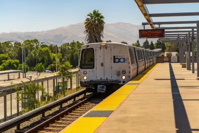 San Francisco Bay Area Rapid Transit train at elevated station with hills and palm tree in background.